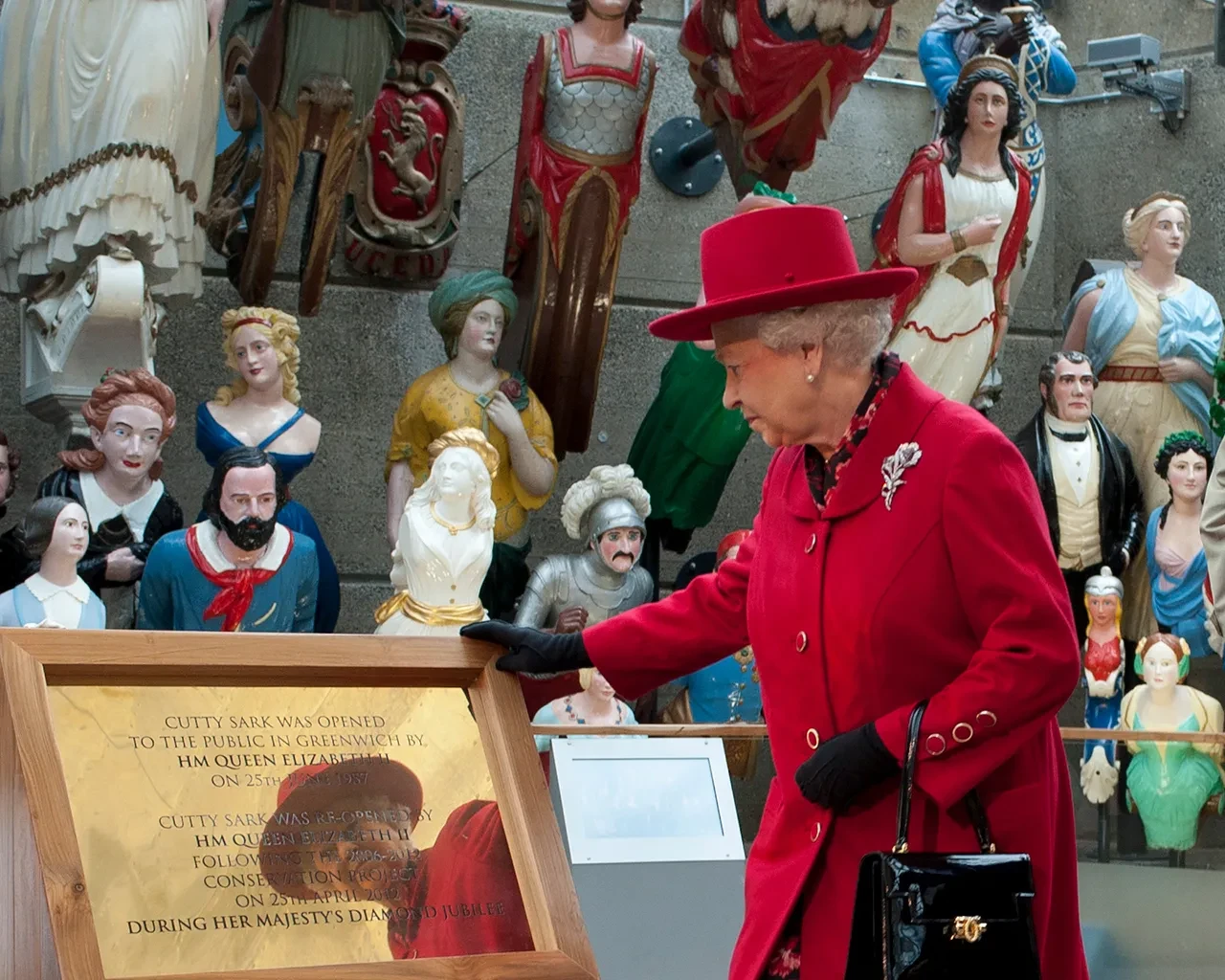 Queen Elizabeth the Second, wearing a red coat and hat, examines a plaque in front of a display of ship's figureheads.